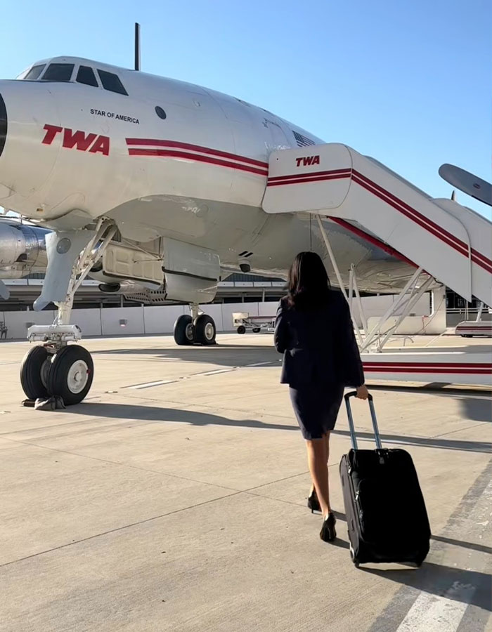 Flight attendant walking with a suitcase toward a vintage TWA airplane on a sunny airport tarmac.