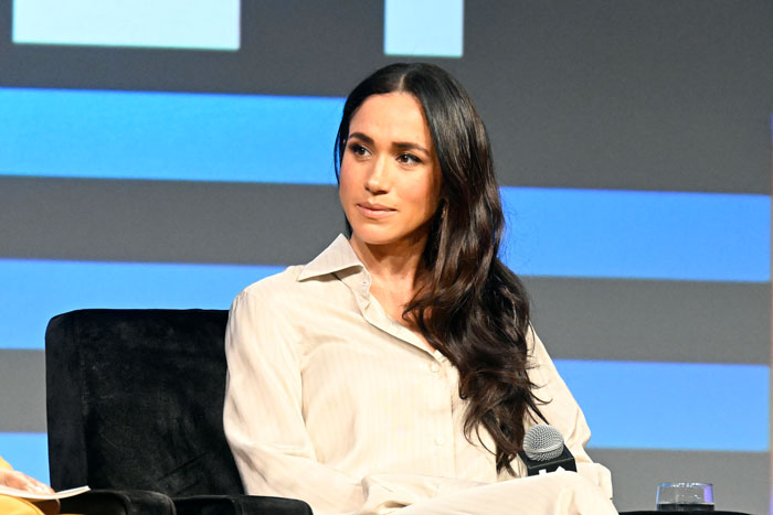 Meghan Markle seated holding a microphone during a panel discussion with a blue and black background.