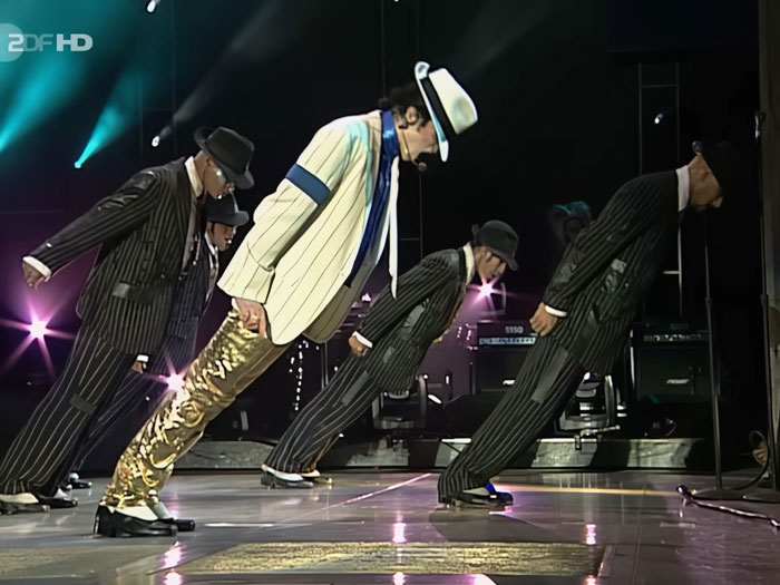 Michael Jackson performing his iconic lean move on stage with backup dancers in pinstripe suits and hats.