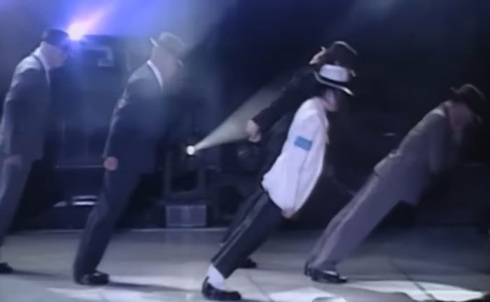 Michael Jackson performing his iconic lean move on stage with backup dancers in suits and fedoras.