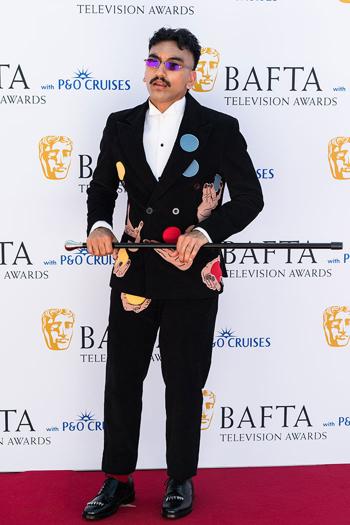 Man wearing a black suit with colorful hand and circle designs, purple glasses at BAFTA TV Awards red carpet.
