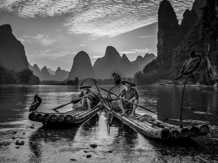 Fishermen on bamboo rafts with birds on a river, framed by karst mountains in a striking 2025 One Shot photo contest image.