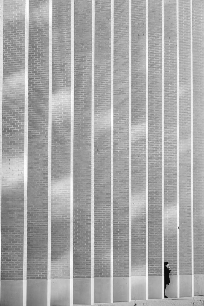 Minimalist black and white photo showing person standing against a tall brick wall with strong vertical lines, one shot photo contest.