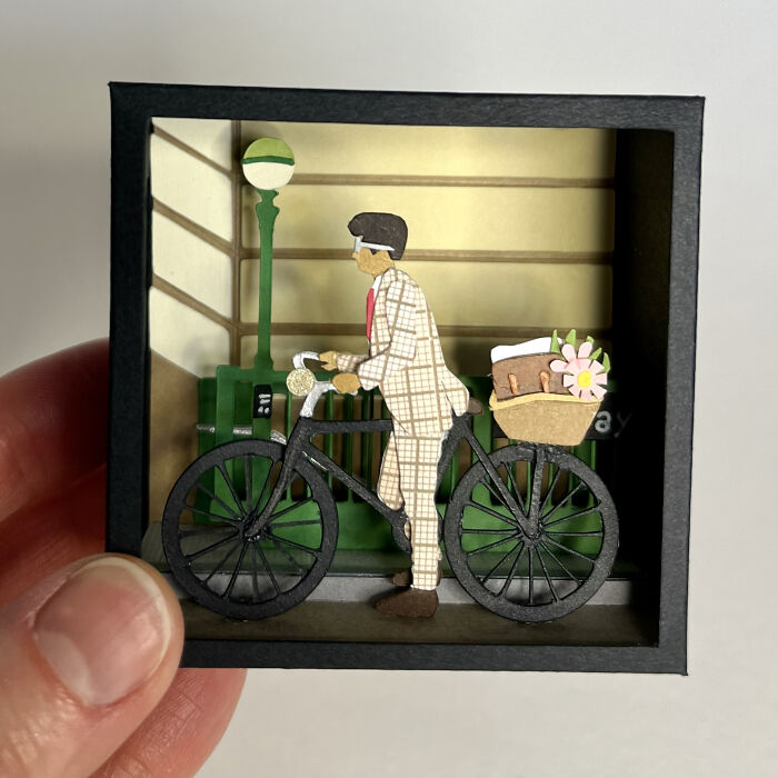Handcrafted intricate colorful paper art of a man with bicycle and basket in a detailed miniature urban scene.