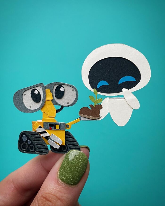 Hand holding intricate colorful paper art of two robots exchanging a small plant against a blue background.