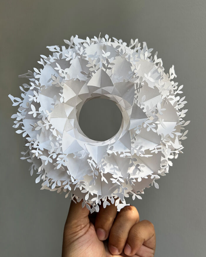 Intricate white paper art sculpture with layered floral and geometric cutouts held by a hand against a neutral background.