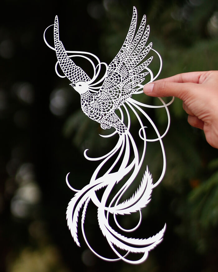 Hand holding an intricate and colorful paper art cutout of a detailed bird with flowing tail feathers against a dark background.