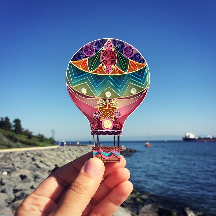 Hand holding an intricate and colorful paper art hot air balloon against a clear blue sky and ocean background.