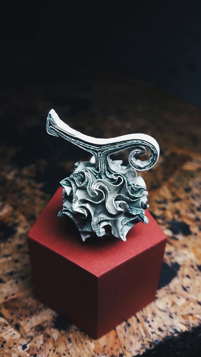 Intricate paper art sculpture with detailed folds and curves displayed on a red cube against a dark background.