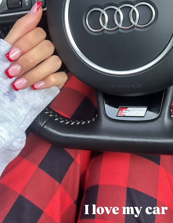 Princess Andre holding steering wheel of Audi S line car wearing red checkered pants and red French manicure nails.