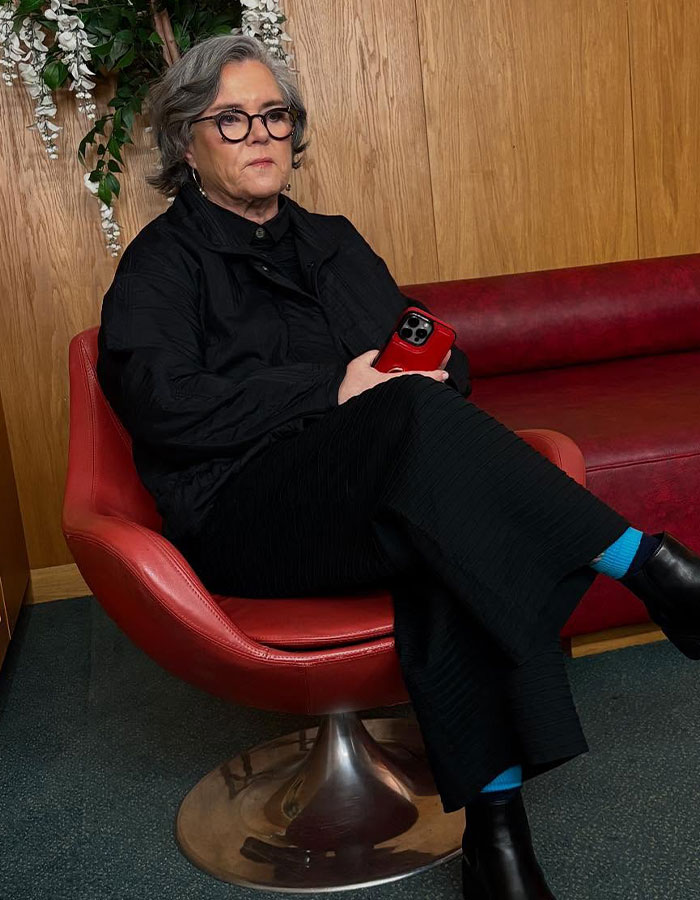 Rosie O&rsquo;Donnell sitting in a red chair indoors, wearing black clothes and holding a red phone, showcasing Mounjaro weight loss.