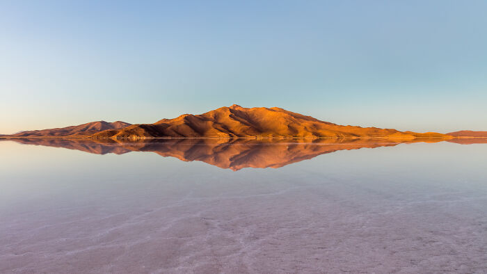 Golden mountain range reflecting on calm salt flat water capturing rare natural phenomena in a serene landscape at sunrise.