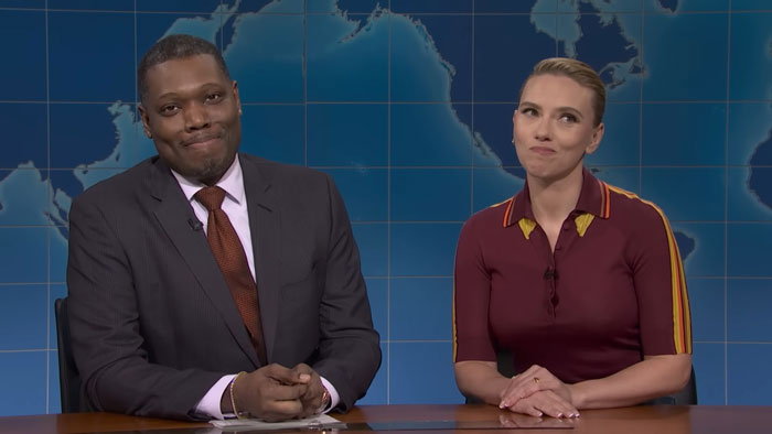 Scarlett Johansson and husband seated at SNL desk during comedy sketch, responding to roast beef joke with smirks. Scarlett Johansson and husband seated at SNL desk during comedy sketch, responding to roast beef joke with smirks.