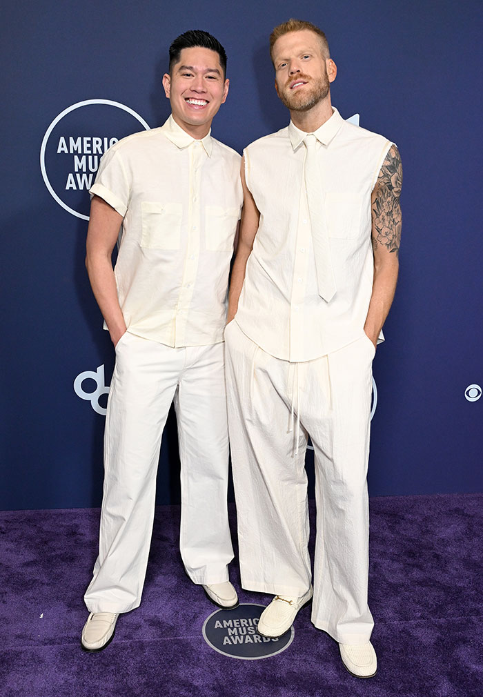 Two men posing at the 2025 American Music Awards wearing matching white sleeveless shirts and wide pants at event carpet.