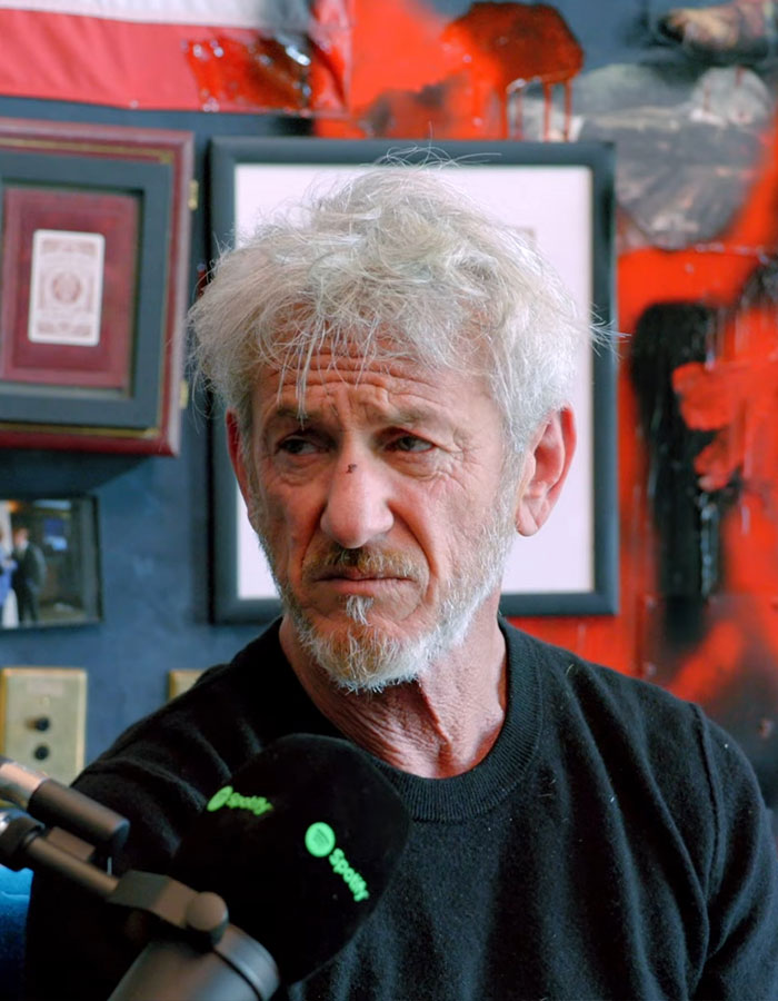 Sean Penn with gray hair and beard, giving a serious expression during a podcast interview indoors.