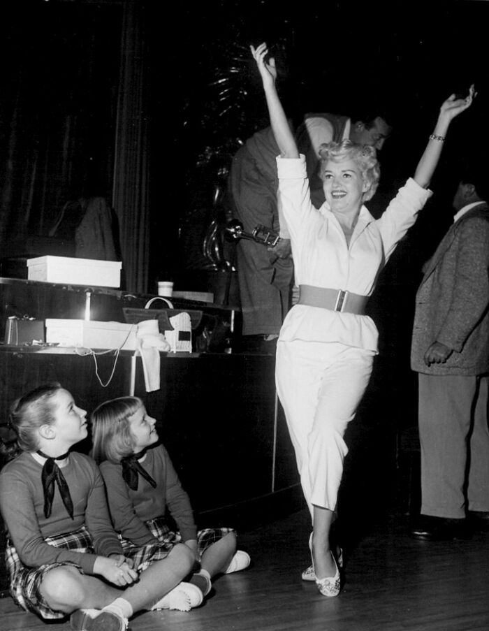 1940s glamorous Hollywood icon in a white outfit joyfully raising arms on stage with two children seated nearby.