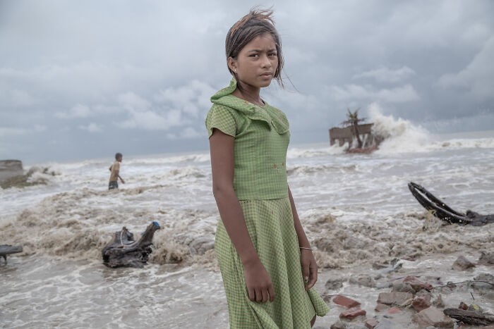 Young girl in green dress standing by turbulent waters and mangrove debris in a powerful mangrove photography awards image.