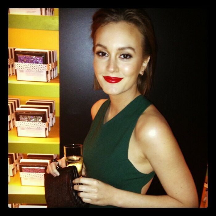 Young woman with red lipstick and green dress holding a clutch and drink in a dimly lit setting, celebrity Instagram post.