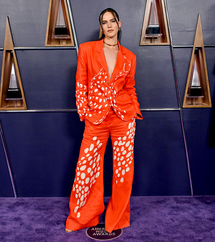 Person wearing a bold orange suit with white spots posing on the purple carpet at the 2025 American Music Awards worst dressed stars event.