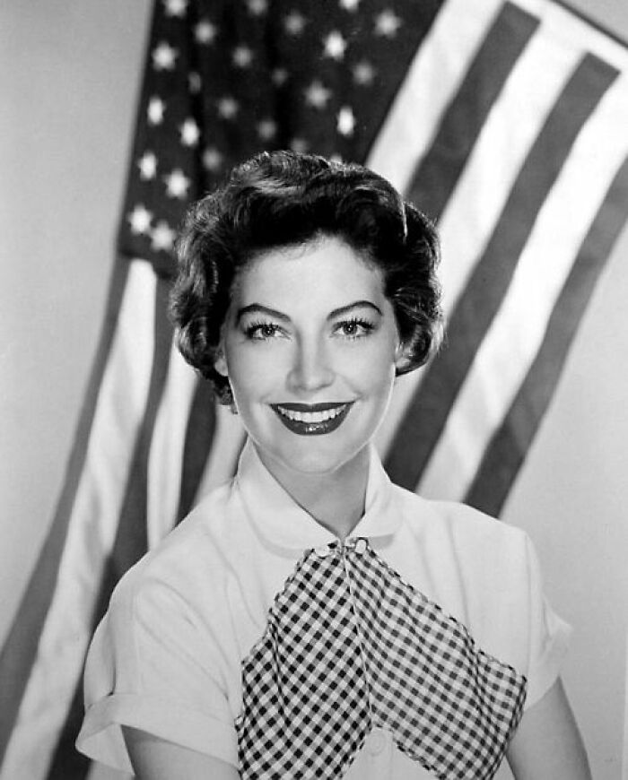 Black and white portrait of a glamorous 1940s Hollywood icon smiling in front of an American flag.