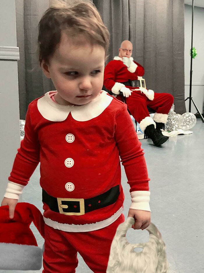 Toddler in a funny Santa outfit holding a hat and beard with a photoshopped Santa in the background showing hilarious Photoshop skills.