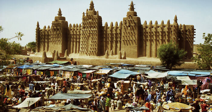 Mud-brick UNESCO treasure in Mali towering over a bustling market filled with people and colorful stalls.