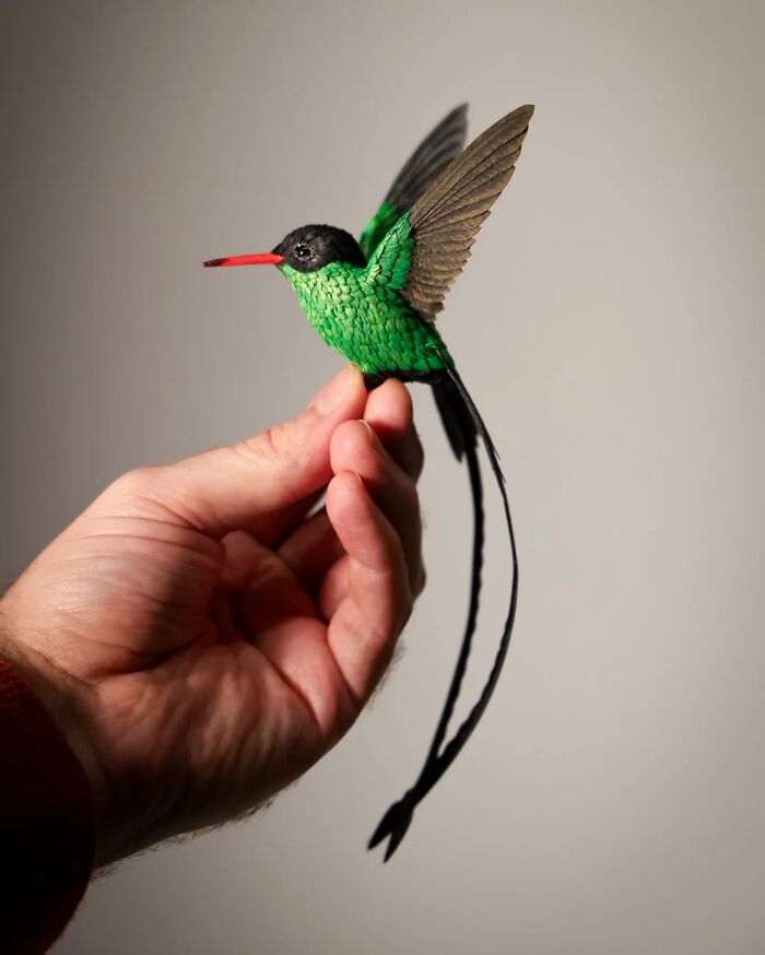 Hand holding a lifelike bird crafted from paper and wood with vibrant green feathers and long tail feathers.