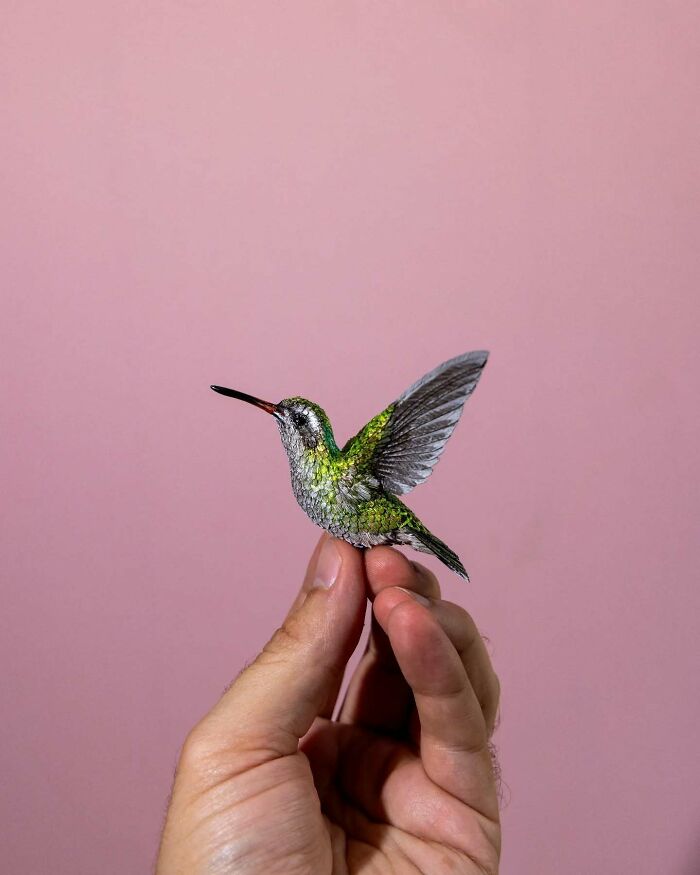 A lifelike bird sculpture made from paper and wood held delicately by a hand against a pink background.