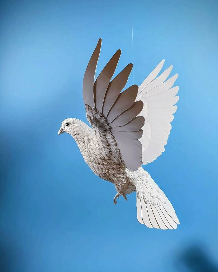 Lifelike bird crafted from paper and wood with detailed feathers, suspended against a blue background.