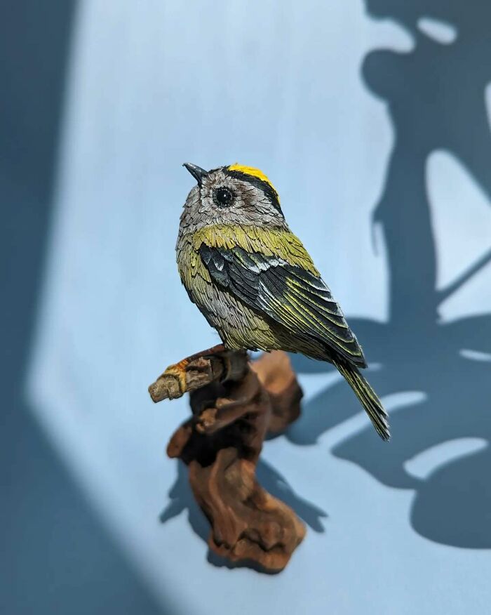 Lifelike bird sculpture crafted from paper and wood, perched on a wooden branch with detailed feathers and vibrant colors.