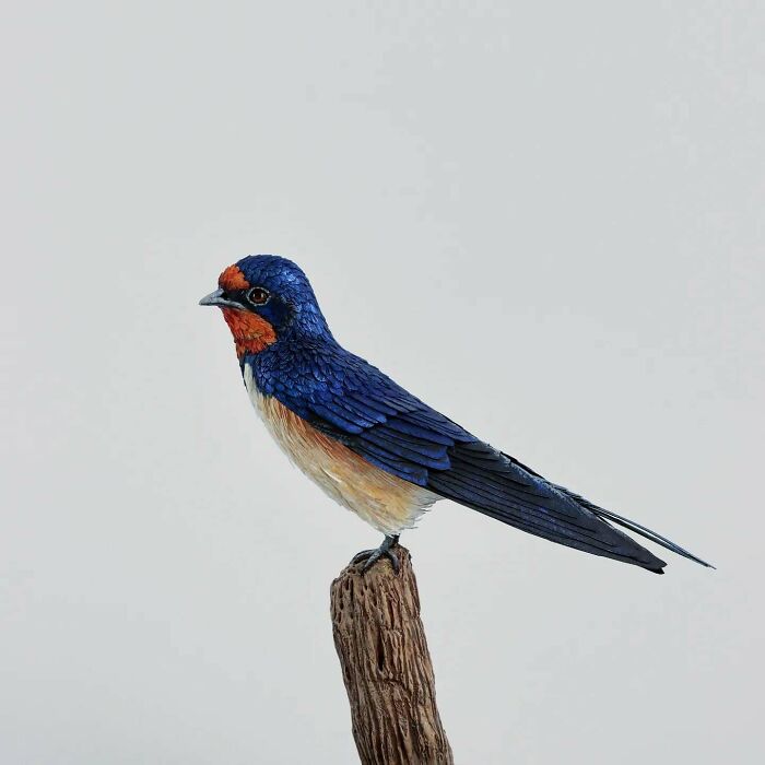 Lifelike bird sculpture crafted from paper and wood, perched on a textured wooden branch against a plain background.