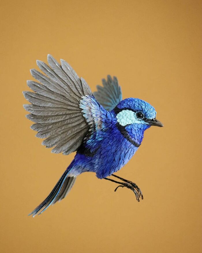 Lifelike blue bird crafted from paper and wood, showcasing intricate feather details and dynamic wing position on a plain background.