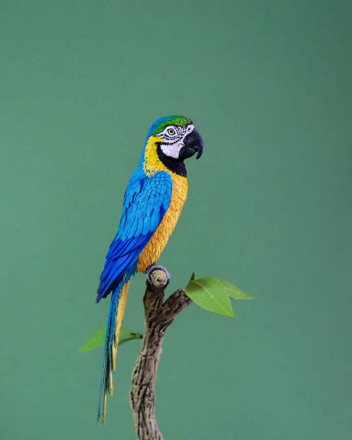 Lifelike bird sculpture crafted from paper and wood, showing detailed feathers and vibrant colors on a branch.