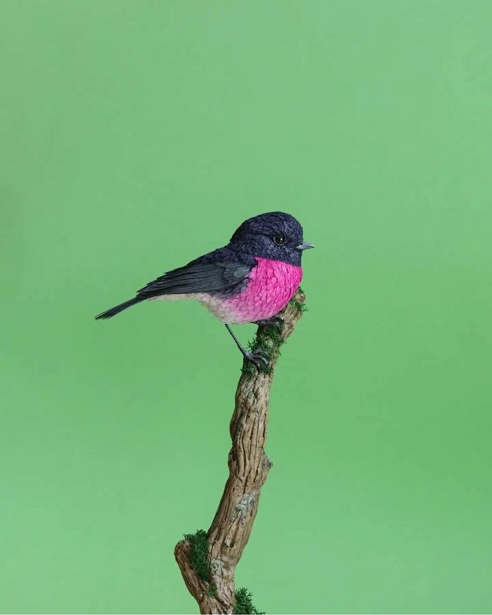 Lifelike bird crafted from paper and wood with vibrant pink and black feathers perched on a textured branch.