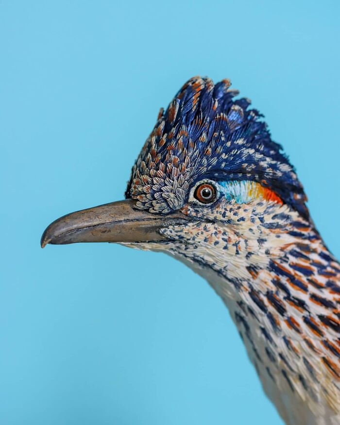 Lifelike bird sculpture crafted from paper and wood, showing detailed feathers and vibrant colors against a blue background.