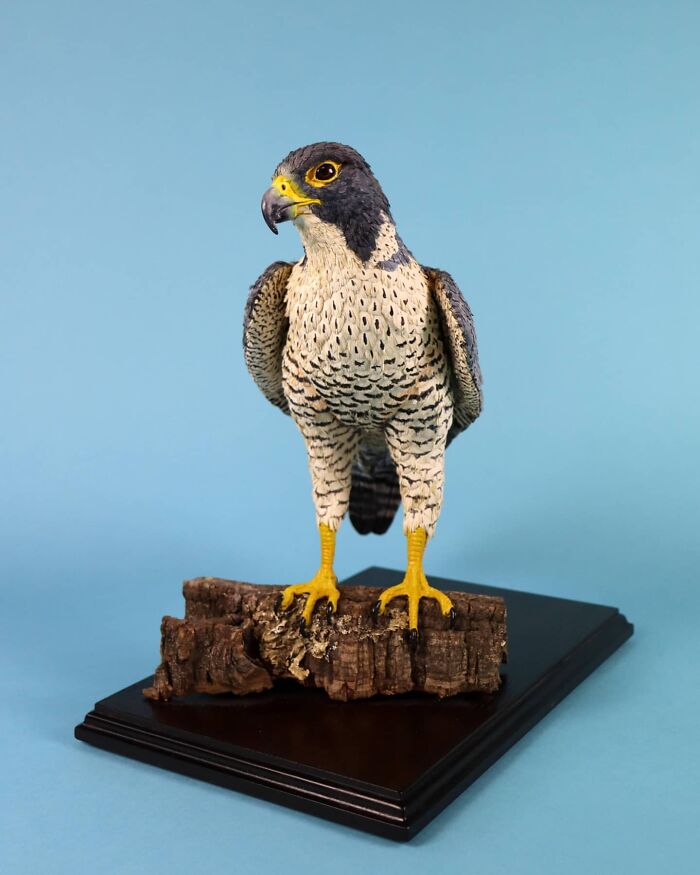 Lifelike bird sculpture crafted from paper and wood, perched on a piece of bark against a blue background.