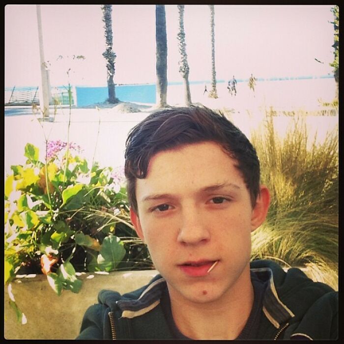 Young celebrity taking a casual selfie outdoors with trees and plants in the background for Instagram posts.