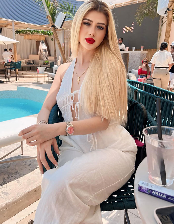 Beauty influencer with long blonde hair and red lipstick sitting poolside at an outdoor cafe during daytime.