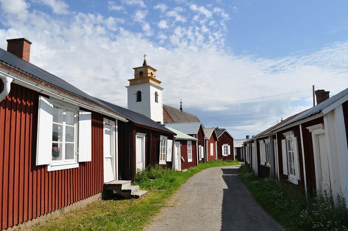 Traditional wooden houses along a path in a hidden UNESCO gem village with a church tower under a partly cloudy sky.