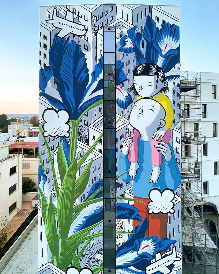 Large mural by Italian artist on city wall depicting two cartoon figures surrounded by oversized blue flowers and city buildings.
