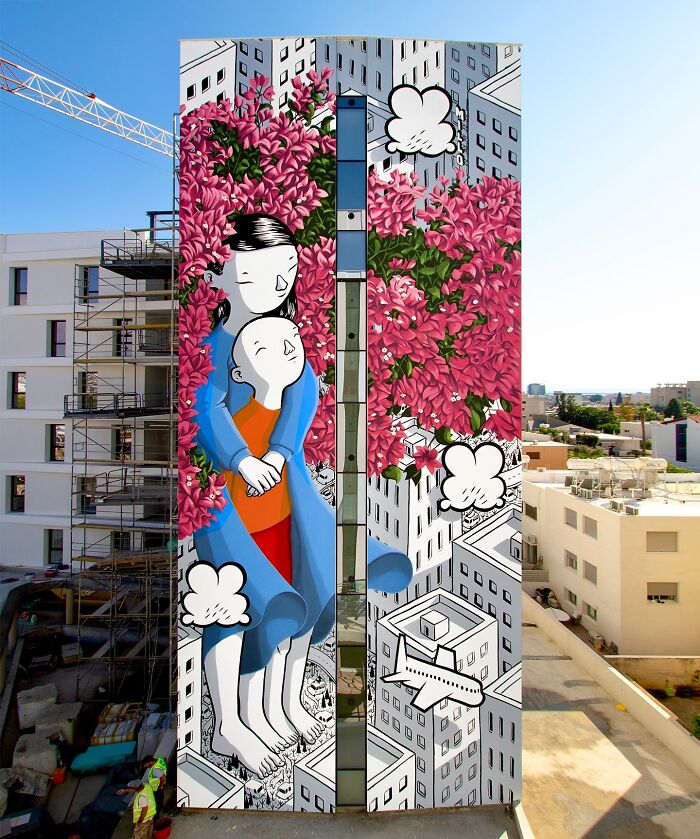 Large mural by Italian artist on city wall, featuring whimsical figures embraced among bright pink flowers and cartoon clouds.