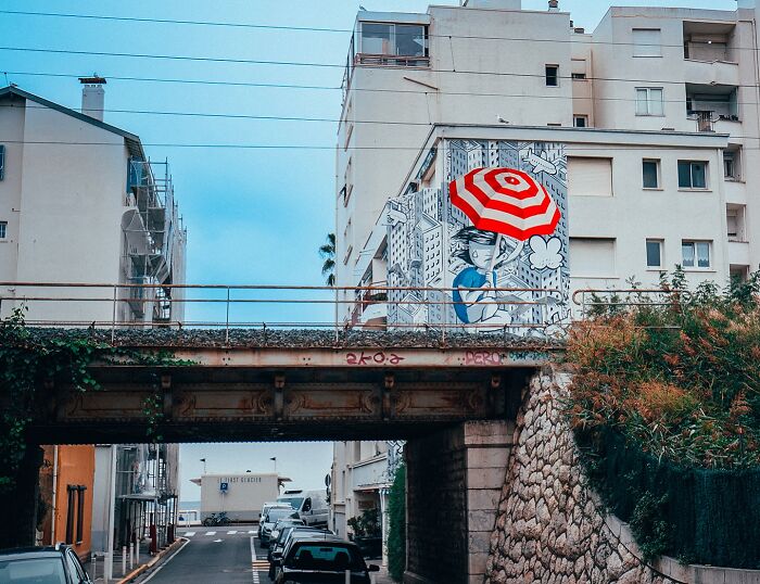 A city wall mural by an Italian artist featuring a whimsical character holding a red and white umbrella in an urban setting.