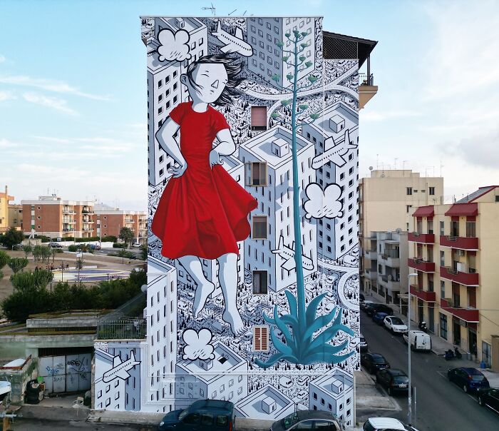 Mural by Italian artist featuring a whimsical girl in a red dress on city walls with cartoon planes and buildings.