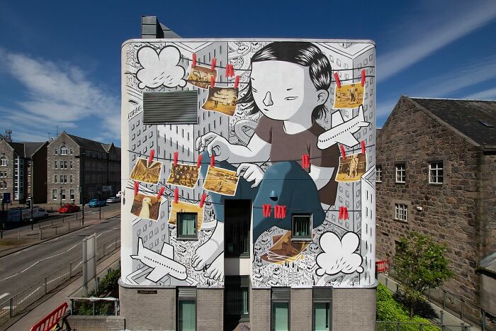 Mural by Italian artist features whimsical city wall art with large child figure and hanging photos in urban setting.