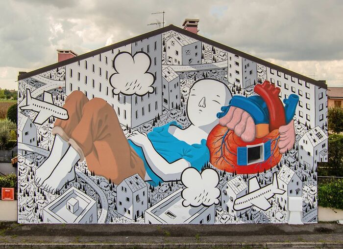Mural by Italian artist showing a person with an anatomical heart amidst cityscape, illustrating whimsical street art on a building wall.