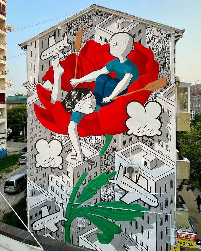 Mural by Italian artist depicting whimsical city walls with oversized flower and characters rowing on red petals.