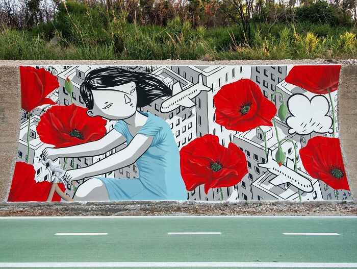 Mural by Italian artist showing whimsical cityscape with a girl on a bicycle and large red flowers on a city wall.