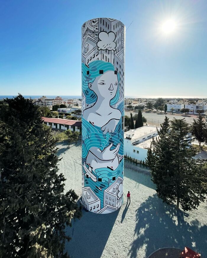 Tall cylindrical structure painted with whimsical city wall mural by Italian artist under clear blue sky.