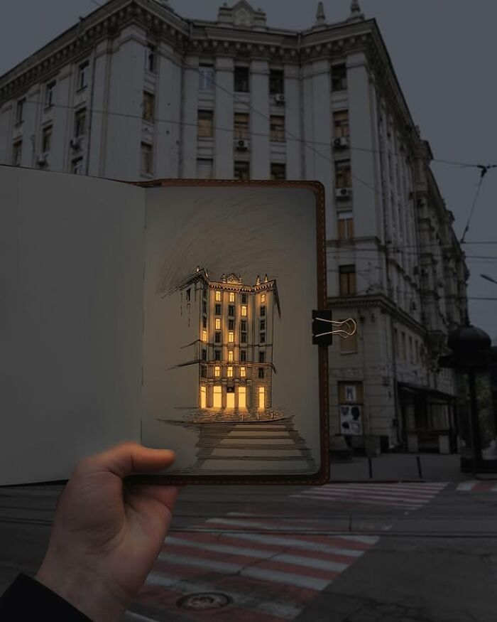 Pen and ink drawing of a building glowing with real light, held up in front of the matching urban background at dusk.
