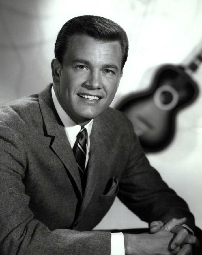 Black and white portrait of a notable person smiling in a suit with a guitar in the background for notable people who passed away.
